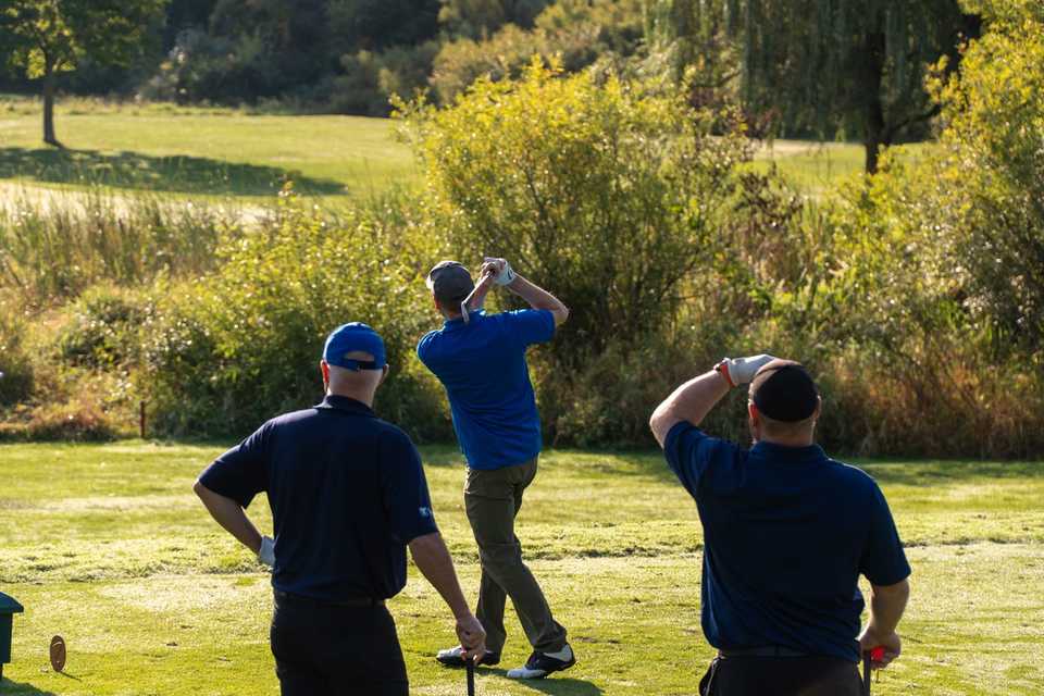 Golfer at the top of a backswing on a tree-lined course in warm late-day light.