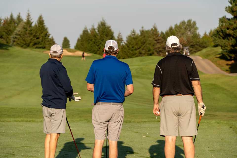 Three golfers on a tee box watching a drive, with fairway and trees in the background.